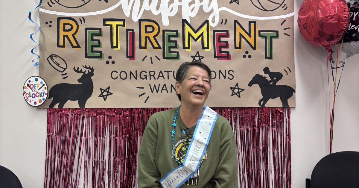 Wanda is seated in front a hand painted poster that reads 'happy retirement' in colourful block font. She is laughing while wearing a white retirement sash.